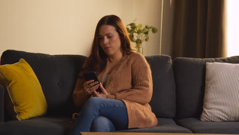 Woman-Sitting-On-Sofa-At-Home-Using-Mobile-Phone-To-Check-Social-Media-Message-And-Read-News-Article